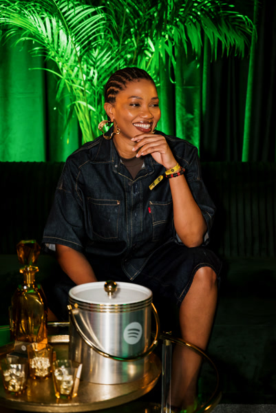 Luedji Luna sorrindo com o queixo apoiado na mão, sentada em um lounge com iluminação verde e plantas tropicais, com um balde de gelo da marca Spotify e um carrinho de bar dourado em primeiro plano