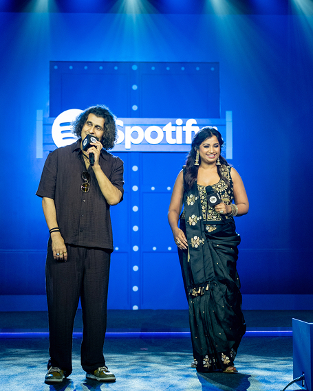 Shreya Goshal and Sonu Nigam on a well-lit stage, with Sonu speaking to the audience