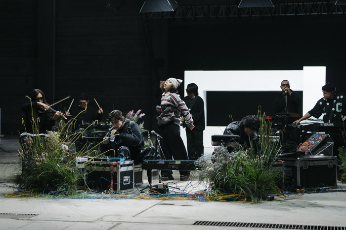 Røz y CoastCity dando una presentación juntos en un escenario decorado con flores, un sintetizador Moog, cables dispersos y un fondo blanco brillante